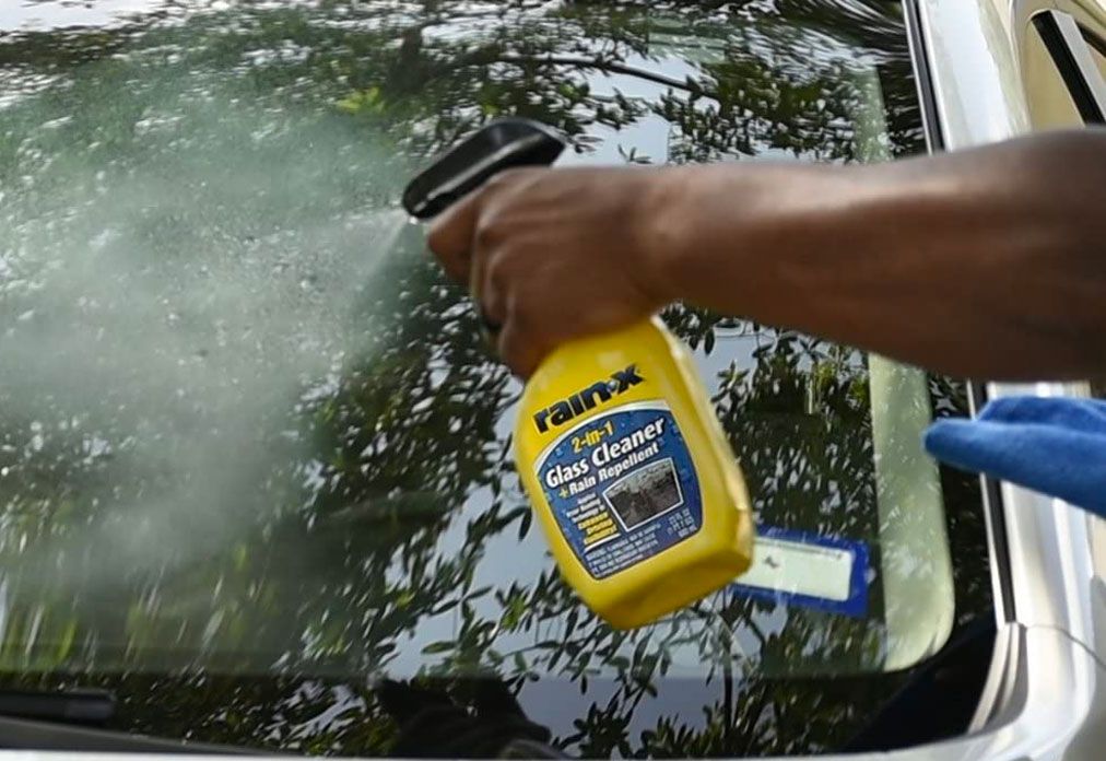 Rain-X 2-in-1 Glass Cleaner + Rain Repellent product image of a yellow spray bottle being applied to a car window.