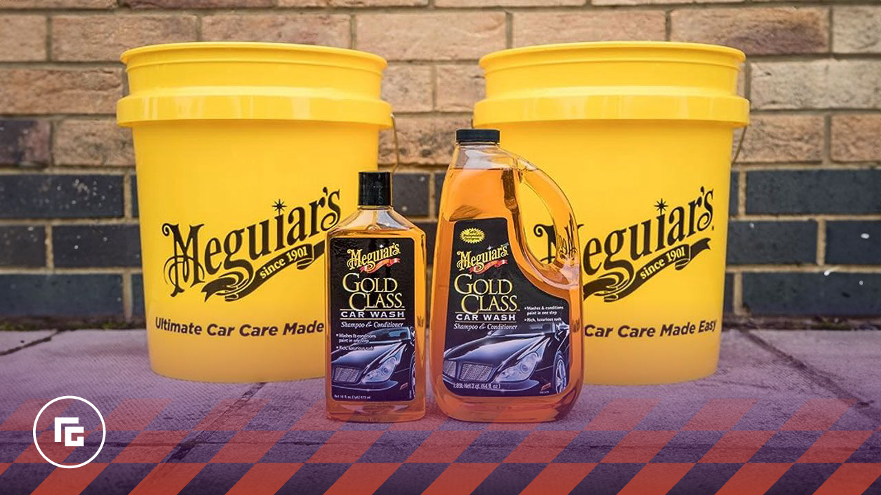 Two bottles containing a golden orange car shampoo in front of two yellow buckets and a brick wall.