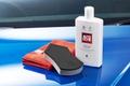 Image of a white bottle of car polish on top of a blue car next to a red cloth and black and grey sponge.