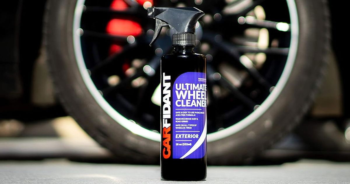 A black spray bottle with blue, red, and white branding on the front in front of a black alloy wheel.