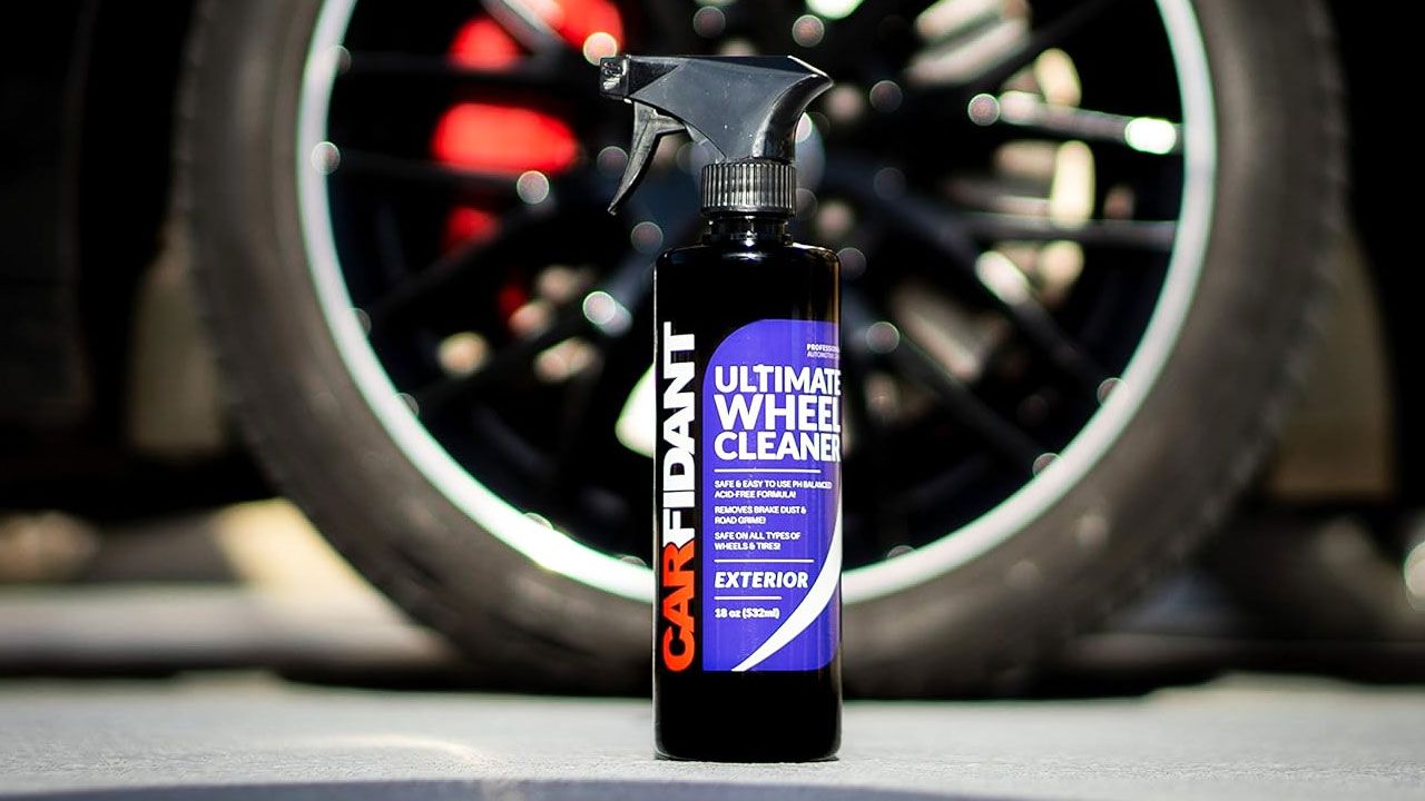 A black spray bottle with blue, red, and white branding on the front in front of a black alloy wheel.