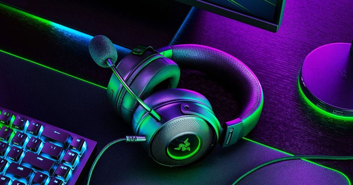 A black wired headset featuring a mic that extends around the front and green Razer branding on the side sat on a desk and bathed in purple light.