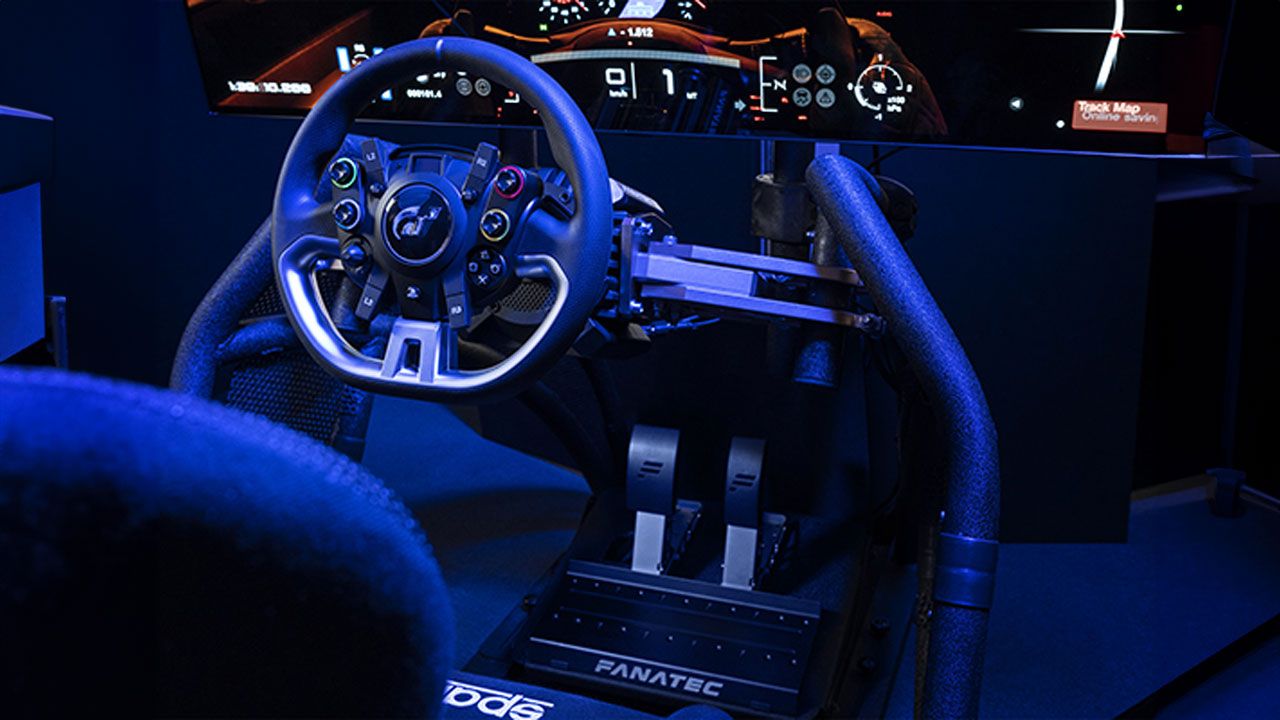 A black racing wheel attached to a rig and TV in front of a racing seat in a darkened room surrounded by blue light.