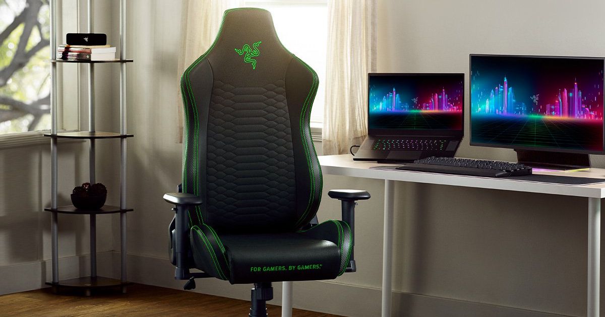 A black office-style gaming chair featuring green trim and Razer branding next to a desk with a laptop, monitor, and keyboard on it.
