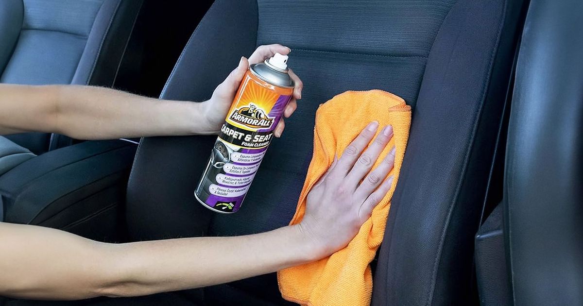 Someone using an orange cloth while spraying a purple and orange can to clean a black car seat.