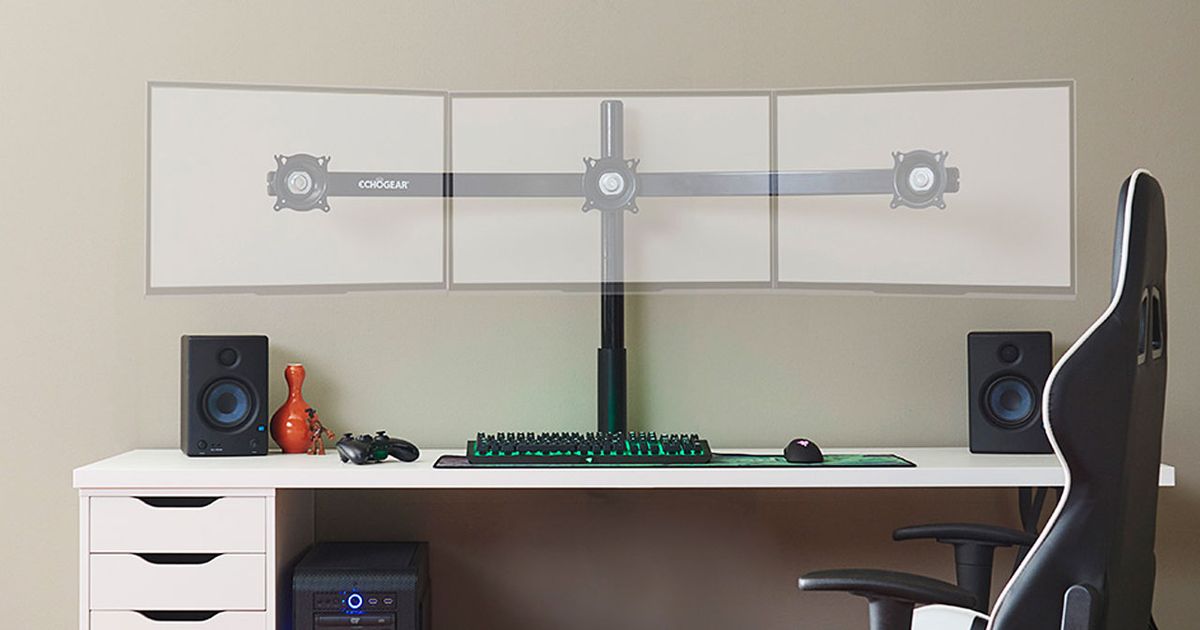 A black triple monitor stand without monitors mounted sat at a desk with a keyboard, mouse, controller, and speaker.