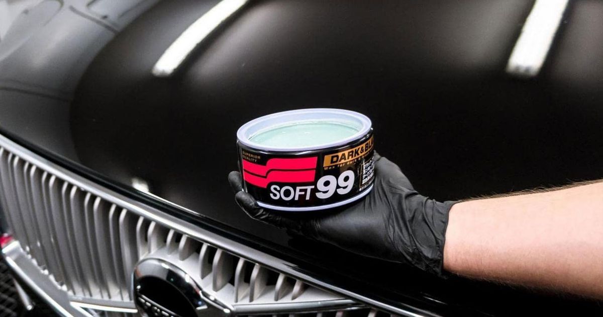 Someone in a black glove holding a black tub of wax featuring red, gold, and white branding in front of a black car.